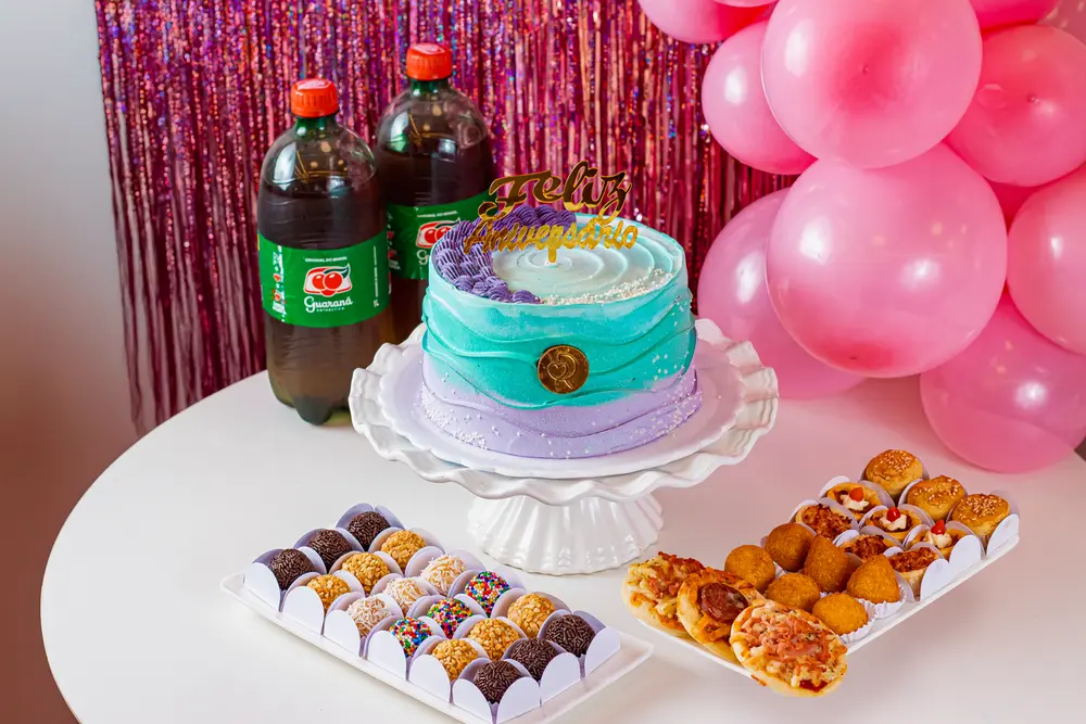 Kit com bolo azul e lilás, docinhos, salgados e guaraná em mesa de festa com balões rosa